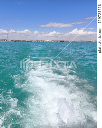 An azure ship's wake on the waves, a panorama of the city on the horizon under a summer sky An azure ship's wake on the waves, a panorama of the city on the horizon under a summer sky 139725518