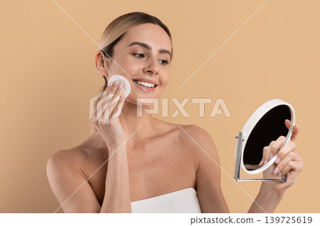 Beautiful smiling woman removing makeup with cotton pad while looking in mirror Beautiful smiling woman removing makeup with cotton pad while looking in mirror 139725619