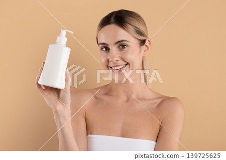 Smiling young woman holding cosmetic lotion bottle and looking at camera Smiling young woman holding cosmetic lotion bottle and looking at camera 139725625