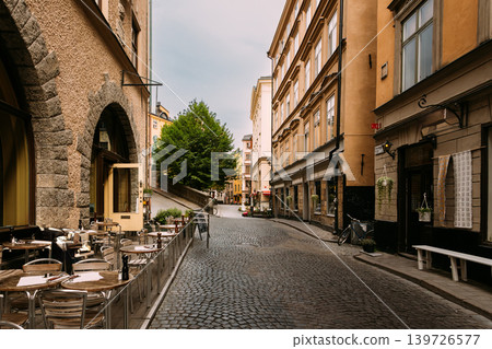 Stone arch passage and outdoor cafe in Gamla Stan, Stockholm, Sweden. Narrow cobblestone street lined with colorful historic buildings and shops in Gamla Stan, Stockholm's old town. Stone arch passage and outdoor cafe in Gamla Stan, Stockholm, Sweden. Narrow cobblestone street lined with colorful historic buildings and shops in Gamla Stan, Stockholm's old town. 139726577