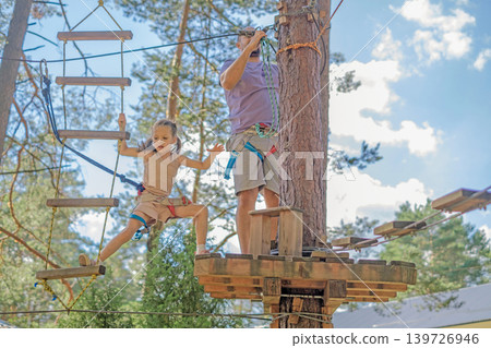 A little girl climbs from a suspended ladder to a step near a tree in a rope aerial town. Passing obstacles on a children's suspension track with my father A little girl climbs from a suspended ladder to a step near a tree in a rope aerial town. Passing obstacles on a children's suspension track with my father 139726946