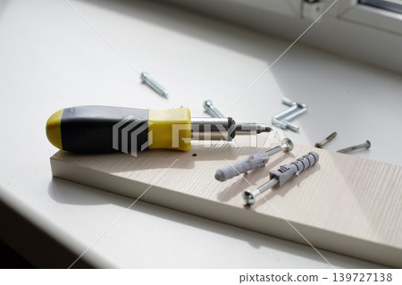 Screwdriver with a black and yellow plastic handle, dowels, and screws on a white window sill on a sunny day. Screwdriver with a black and yellow plastic handle, dowels, and screws on a white window sill on a sunny day. 139727138