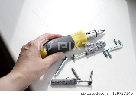 Hand picks up a screwdriver with a black and yellow rubber handle from a white windowsill. Dowels and screws are on the windowsill on a sunny day. Hand picks up a screwdriver with a black and yellow rubber handle from a white windowsill. Dowels and screws are on the windowsill on a sunny day. 139727140