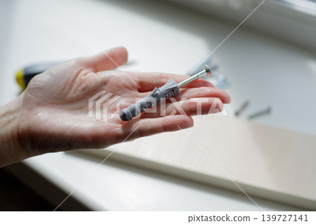 Hand holds a gray plastic dowel against a white windowsill. Dowels and screws are scattered on the windowsill on a sunny day. Hand holds a gray plastic dowel against a white windowsill. Dowels and screws are scattered on the windowsill on a sunny day. 139727141