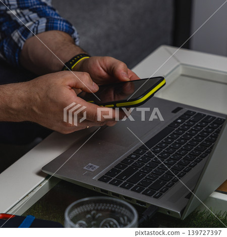 Man holding smartphone while working on laptop remotely Man holding smartphone while working on laptop remotely 139727397