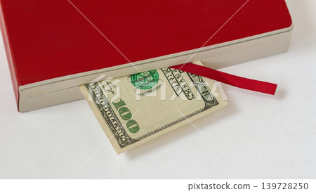 Money hidden under red notebook shows one hundred dollar bill partly covered by book pages and ribbon bookmark on white surface. Concept of money, savings, finance, cost, debt, payment Money hidden under red notebook shows one hundred dollar bill partly covered by book pages and ribbon bookmark on white surface. Concept of money, savings, finance, cost, debt, payment 139728250