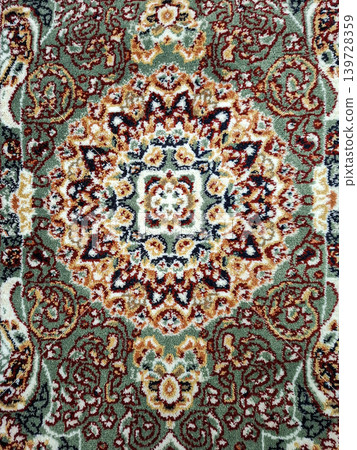 This carpet features a top view of a traditional oriental design. The detailed pattern includes green, orange, and red colors arranged in ornate shapes. This carpet features a top view of a traditional oriental design. The detailed pattern includes green, orange, and red colors arranged in ornate shapes. 139728359
