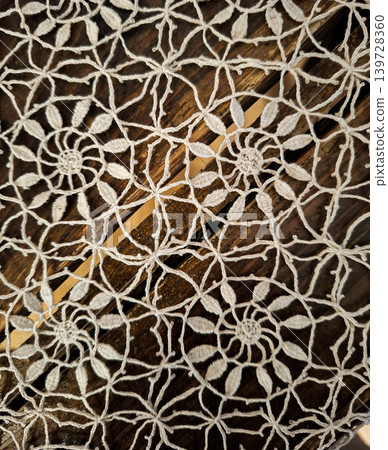 A white crocheted lace tablecloth is laid over a dark wooden table. The tablecloth features floral patterns and adds charm to the dining area. It is daytime, and natural light enhances the setting. 139728360