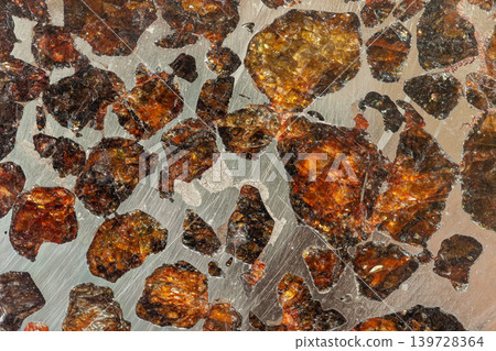 Close-up of polished pallasite meteorite slice, translucent amber olivine crystals embedded in metallic nickel iron matrix. Natural mineral texture for geology, science, space themed design 139728364