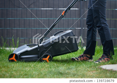 Person stands beside lawn mower on backyard grass near fence Person stands beside lawn mower on backyard grass near fence 139728679