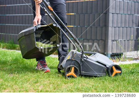 Person lifts grass collector with cut grass from lawn mower in backyard Person lifts grass collector with cut grass from lawn mower in backyard 139728680