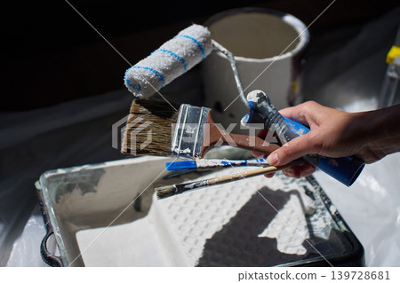 Person holds paint roller and brushes over tray beside paint bucket on protective plastic sheet 139728681