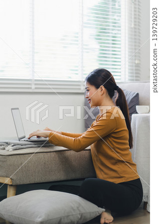 A woman comfortably working on her laptop from home, sitting on the floor in a relaxed and cozy environment 139729203