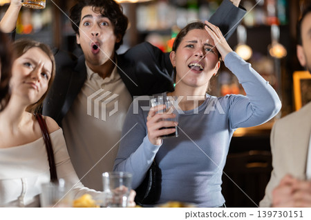 Emotional sad group of young sports fans man and woman worrying about defeat sports team, watching match on TV during rest in beer bar Emotional sad group of young sports fans man and woman worrying about defeat sports team, watching match on TV during rest in beer bar 139730151