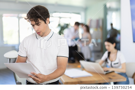 Young man standing in office holds paper documents Young man standing in office holds paper documents 139730254