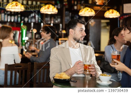 Couple of young guy and girl chatting, drinking beer and eating snacks at table in sports pub Couple of young guy and girl chatting, drinking beer and eating snacks at table in sports pub 139730439