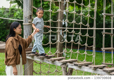 preschool child girl walking on a wooden balancing bridge with her mother at outdoor playground 139730608