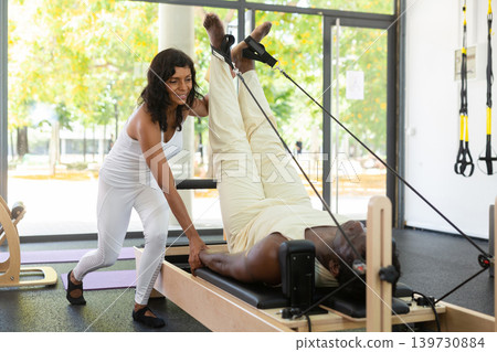 Focused hispanic instructor helping African American man practicing pilates. Individual coach Focused hispanic instructor helping African American man practicing pilates. Individual coach 139730884