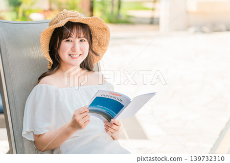 年輕的亞洲女性坐在泳池邊的躺椅上閱讀旅遊指南 年輕的亞洲女性坐在泳池邊的躺椅上閱讀旅遊指南 139732310
