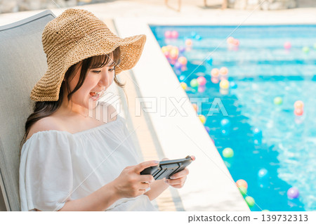 年輕的亞洲女性坐在泳池邊的躺椅上玩遊戲 年輕的亞洲女性坐在泳池邊的躺椅上玩遊戲 139732313