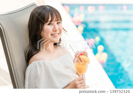 年輕的亞洲女性坐在泳池邊的躺椅上喝熱帶果汁 年輕的亞洲女性坐在泳池邊的躺椅上喝熱帶果汁 139732320