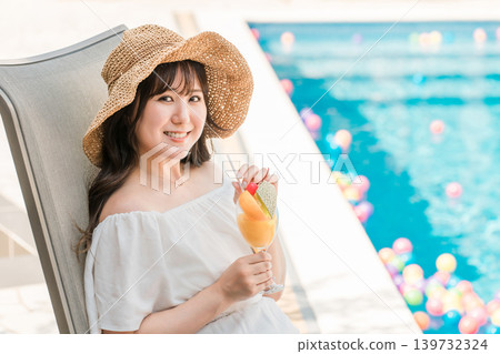 Young Asian woman sitting on a recliner by the pool and drinking tropical juice Young Asian woman sitting on a recliner by the pool and drinking tropical juice 139732324