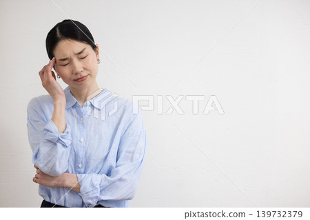 A Japanese woman stands in front of a white wall, holding her hand to her temple and making a pained expression. A Japanese woman stands in front of a white wall, holding her hand to her temple and making a pained expression. 139732379