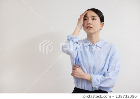 A Japanese woman stands in front of a white wall, holding her hand to her temple and making a pained expression. A Japanese woman stands in front of a white wall, holding her hand to her temple and making a pained expression. 139732380
