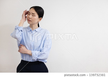 一位日本女子站在白牆前，手按著太陽穴，臉上露出痛苦的表情。 139732381