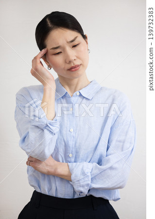A Japanese woman stands in front of a white wall, holding her hand to her temple and making a pained expression. A Japanese woman stands in front of a white wall, holding her hand to her temple and making a pained expression. 139732383