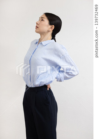 A Japanese woman stands in front of a white wall, hands on her hips, with a pained expression on her face. A Japanese woman stands in front of a white wall, hands on her hips, with a pained expression on her face. 139732469