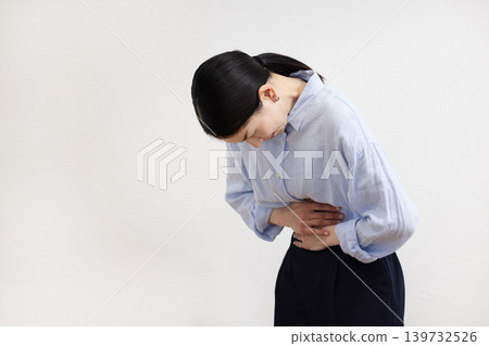 一名日本女子站在白牆前，一手放在肚子上，臉上露出痛苦的表情。 139732526
