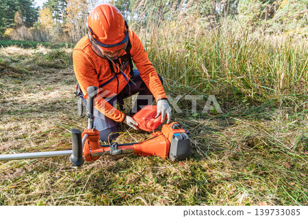 Man refuels brush cutter in field, wearing safety gear for landscaping maintenance work 139733085