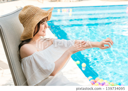 一位年輕的亞洲女性坐在泳池邊的躺椅上,在手臂上塗抹防曬霜 一位年輕的亞洲女性坐在泳池邊的躺椅上,在手臂上塗抹防曬霜 139734095