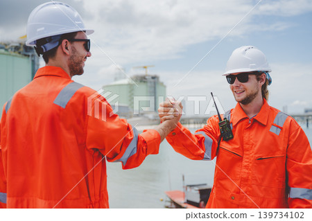 Two engineers in orange protective uniforms shaking hands at LNG terminal near the water 139734102