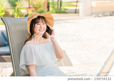 一位年輕的亞洲女性坐在泳池邊的躺椅上,用智慧型手機通話(預訂、搜尋、客房服務) 一位年輕的亞洲女性坐在泳池邊的躺椅上,用智慧型手機通話(預訂、搜尋、客房服務) 139734132