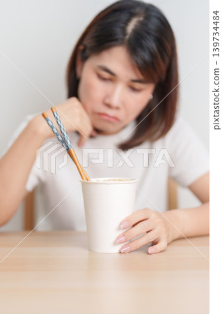 Bored young Asian woman eating instant noodles at home. Exhausted female looking unhappy while eating cup ramen. quick meal after work, burnout and unhealthy eating habit, lifestyle and cheap meal Bored young Asian woman eating instant noodles at home. Exhausted female looking unhappy while eating cup ramen. quick meal after work, burnout and unhealthy eating habit, lifestyle and cheap meal 139734484