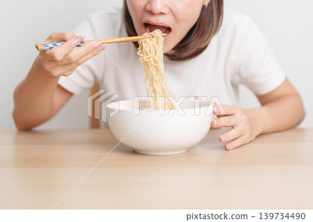 Happy young Asian woman enjoying a delicious bowl of hot instant noodles while relaxing at home, Cheerful female eating instant ramen with a smile at apartment. Simple lifestyle and quick meal concept 139734490