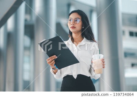 Professional woman in eyeglasses holding iced coffee and a clipboard in a bright office setting, representing calm confidence, corporate lifestyle, waiting time, and everyday work routine. Professional woman in eyeglasses holding iced coffee and a clipboard in a bright office setting, representing calm confidence, corporate lifestyle, waiting time, and everyday work routine. 139734864