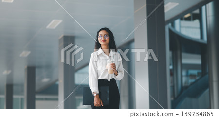 Professional woman in eyeglasses holding iced coffee and a clipboard in a modern office corridor, expressing confident corporate lifestyle, work break, career focus, and everyday productivity. Professional woman in eyeglasses holding iced coffee and a clipboard in a modern office corridor, expressing confident corporate lifestyle, work break, career focus, and everyday productivity. 139734865
