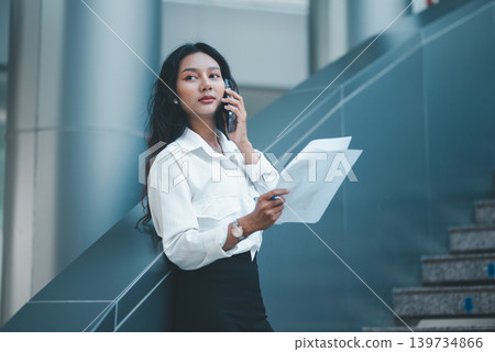 Professional woman balancing a smartphone on her shoulder while reviewing reports, showing multitasking, mobile communication, document checking, office efficiency, and problem solving. Professional woman balancing a smartphone on her shoulder while reviewing reports, showing multitasking, mobile communication, document checking, office efficiency, and problem solving. 139734866