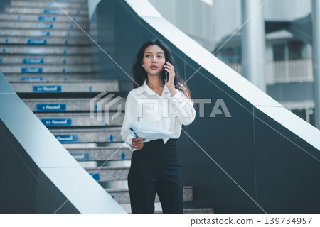 Professional businesswoman talking on a smartphone while holding reports near office stairs, representing mobile communication, client coordination, multitasking, and corporate workflow. Professional businesswoman talking on a smartphone while holding reports near office stairs, representing mobile communication, client coordination, multitasking, and corporate workflow. 139734957
