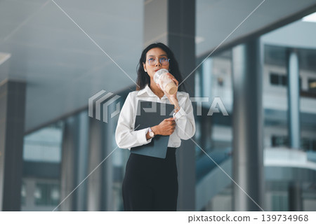 Professional woman in eyeglasses drinking iced coffee while holding a clipboard in a modern office walkway, showing office break, calm confidence, business lifestyle, and daily productivity. 139734968