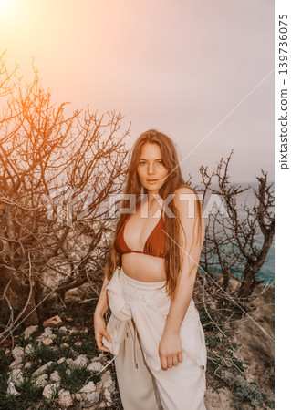 Woman Coast Bikini beautiful young woman in brown bikini top posing near dry bushes on a rocky cliff during golden hour 139736075