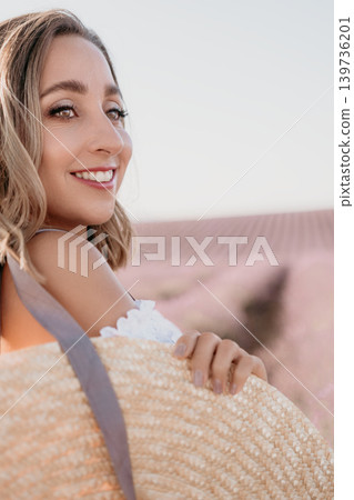 Woman flower field smile looking back over her shoulder while holding a straw hat in a blooming summer meadow Woman flower field smile looking back over her shoulder while holding a straw hat in a blooming summer meadow 139736201