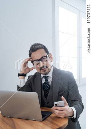 一位中年男性商務人士在使用智慧型手機和筆記型電腦時遇到問題,正在努力解決。 一位中年男性商務人士在使用智慧型手機和筆記型電腦時遇到問題,正在努力解決。 139737351