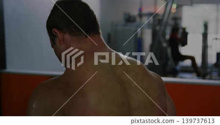 Standing shirtless man flexing back muscles at gym, with mirrored wall and weight machine 139737613