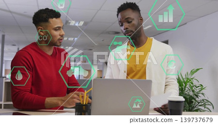 Discussing coworkers in red and cardigan at open-plan office leaning over laptop with data overlays Discussing coworkers in red and cardigan at open-plan office leaning over laptop with data overlays 139737679