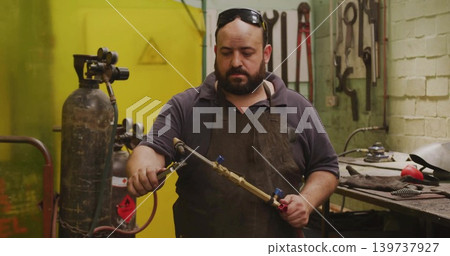 Inspecting bearded welder in dark apron checking oxy-fuel torch valves in shop, with gas cylinders Inspecting bearded welder in dark apron checking oxy-fuel torch valves in shop, with gas cylinders 139737927