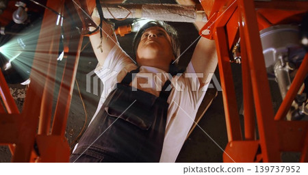 Working man wearing white tee overalls under vehicle at workshop with red frame wiring bolts lamp Working man wearing white tee overalls under vehicle at workshop with red frame wiring bolts lamp 139737952